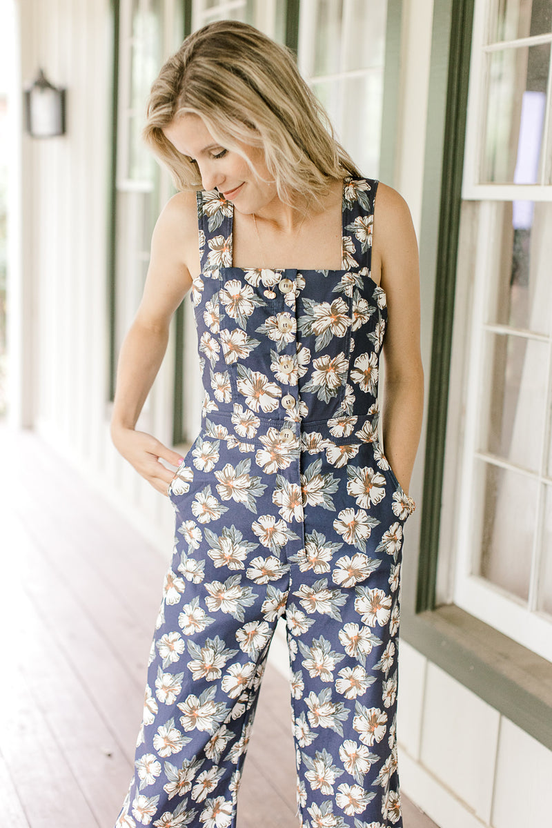 Model wearing a navy jumpsuit with white flowers, button front and pockets. 