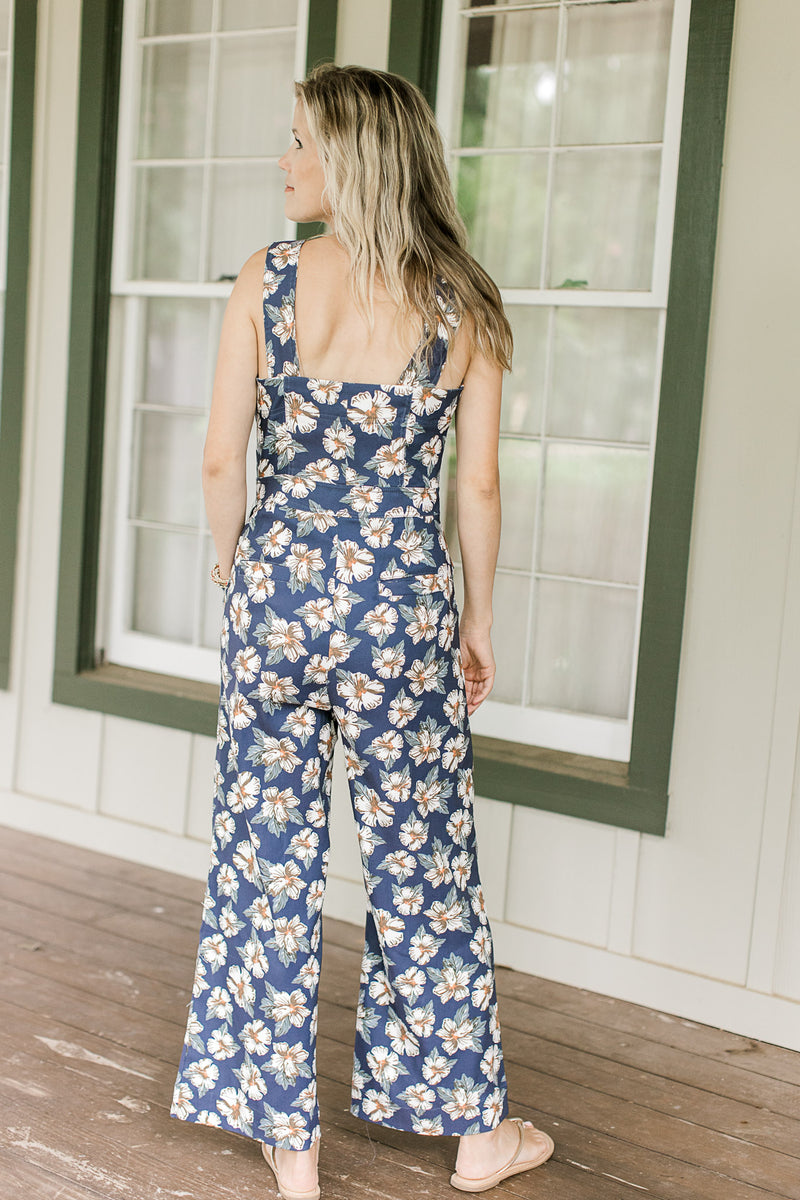 Back view of Model wearing a navy jumpsuit with white flowers, square neck and wide legs.