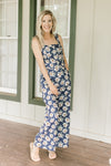 Model wearing sandals with a navy jumpsuit with white flowers, tank sleeves and wide legs.