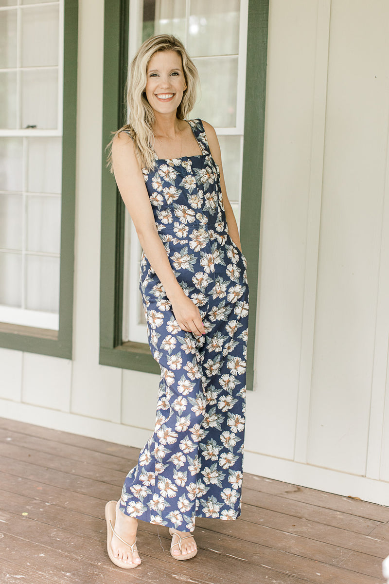 Model wearing sandals with a navy jumpsuit with white flowers, tank sleeves and wide legs.