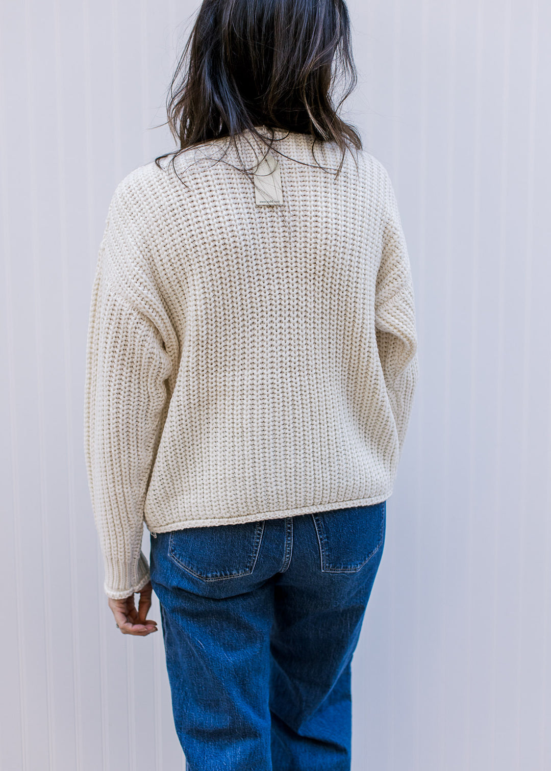 Back view of Model wearing a cream cable knit cardigan with long sleeves and patch pockets. 