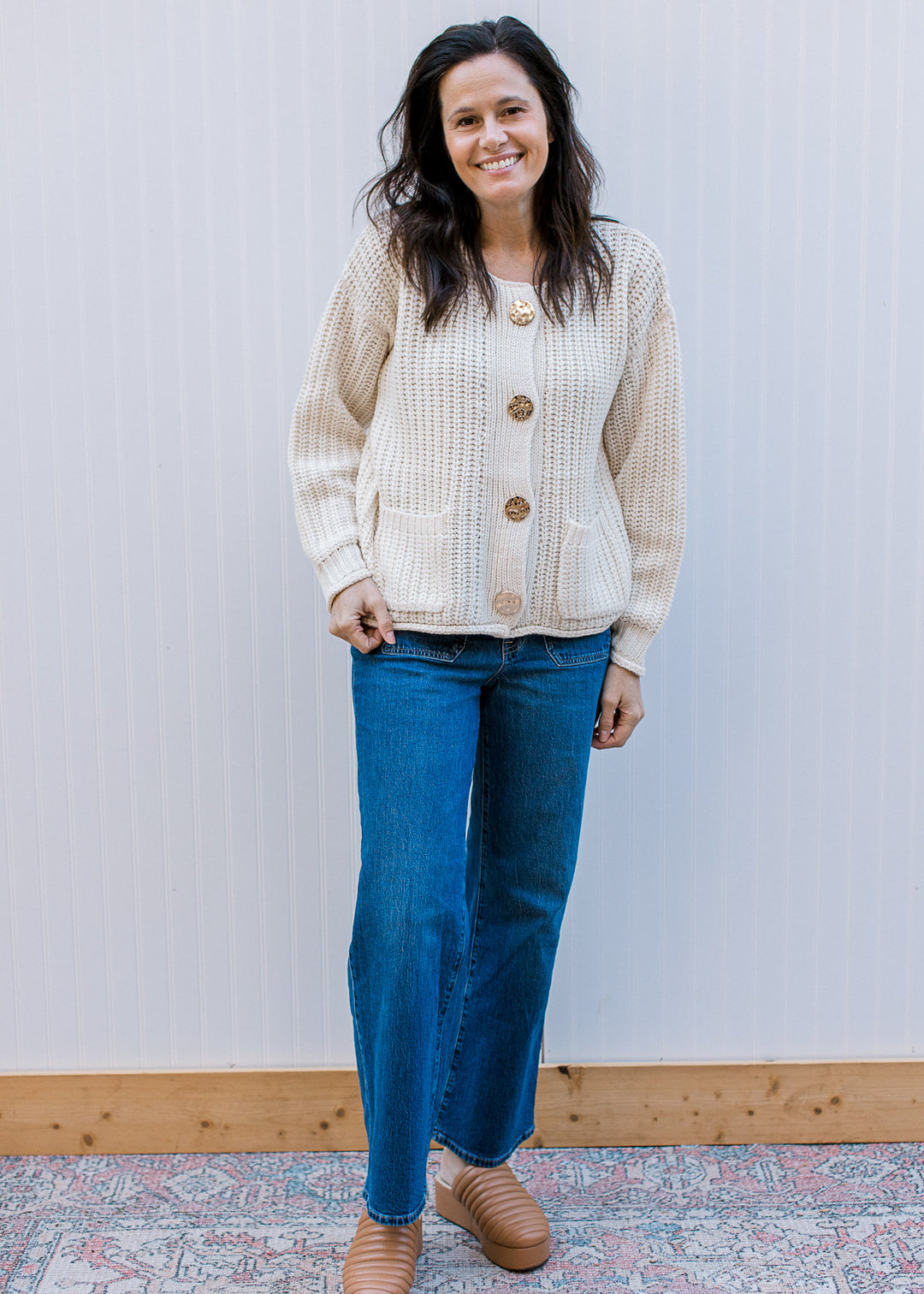 Model wearing jeans, mules and a cream cable knit cardigan, with gold buttons and round neck. 