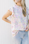 Model wearing a white top with bright painted flowers a mock neck and flutter cap sleeves.