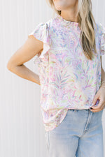 Model wearing a white top with bright painted flowers a mock neck and flutter cap sleeves.