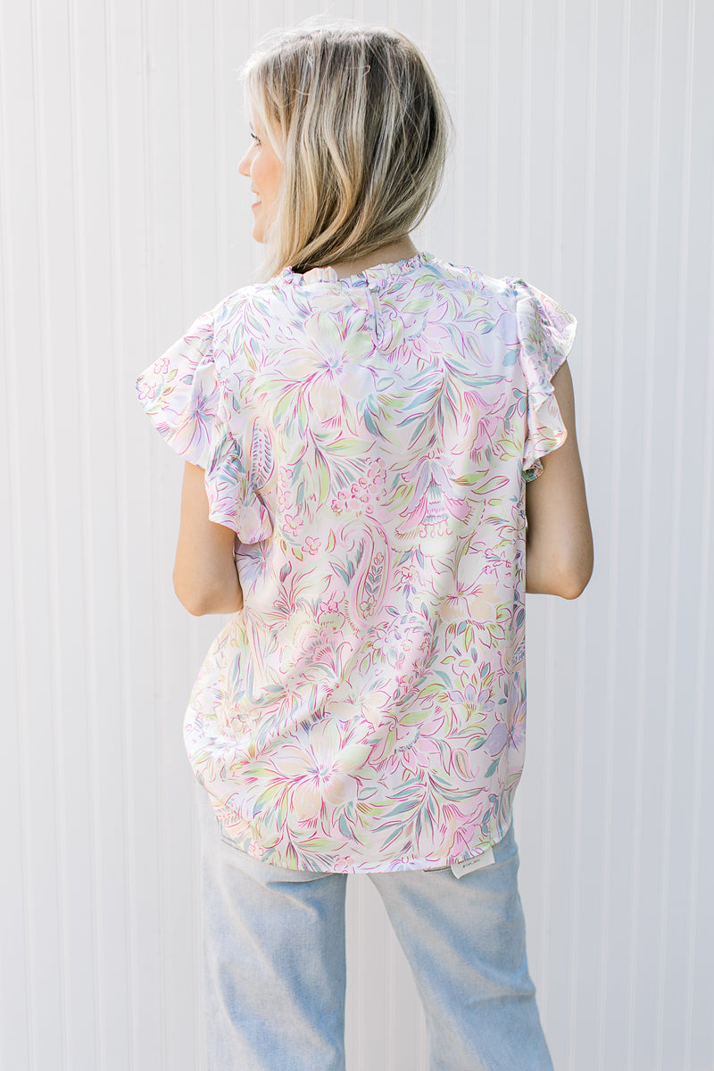 Back view of Model wearing a white top with bright painted flowers and flutter cap sleeves.