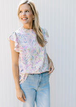 Model wearing jeans with a white top with bright painted flowers and flutter cap sleeves.