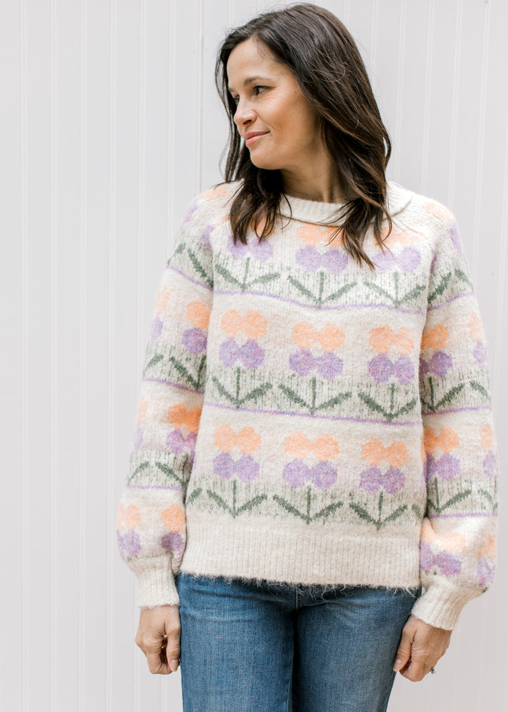 Model wearing a cream sweater with purple and peach flowers, a round neck and long sleeves. 