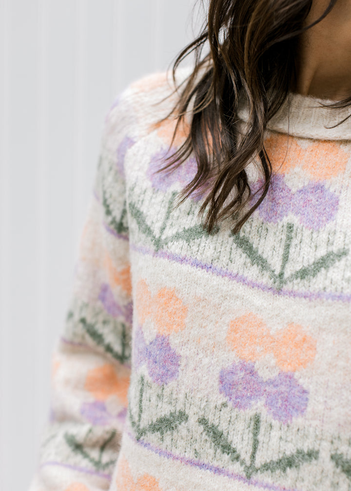 Close up of peach and lavender flowers on a cream sweater with a round neck and long sleeves. 