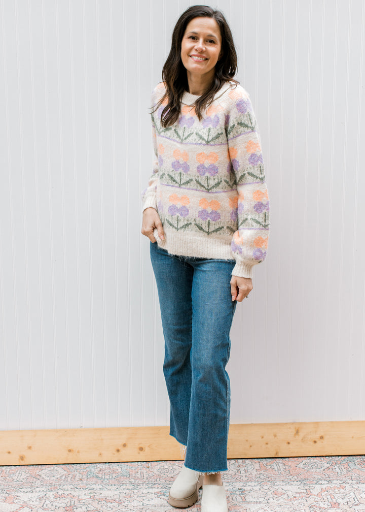 Model wearing jeans and a cream sweater with purple and peach flowers and long sleeves. 