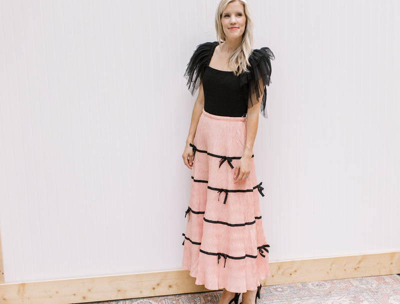 Model wearing a black top with a peach skirt with plisse bows, elastic waist and pleated fabric.