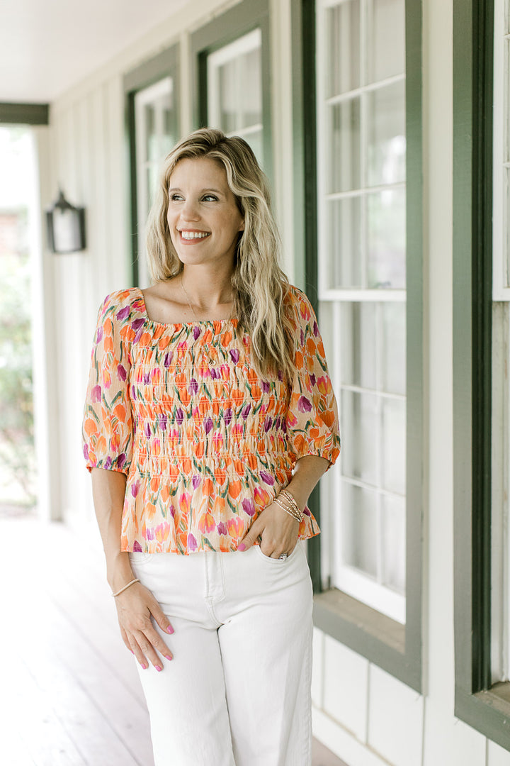 Model wearing white pants and a peach top with purple, gold and green tulips and a smocked bodice.