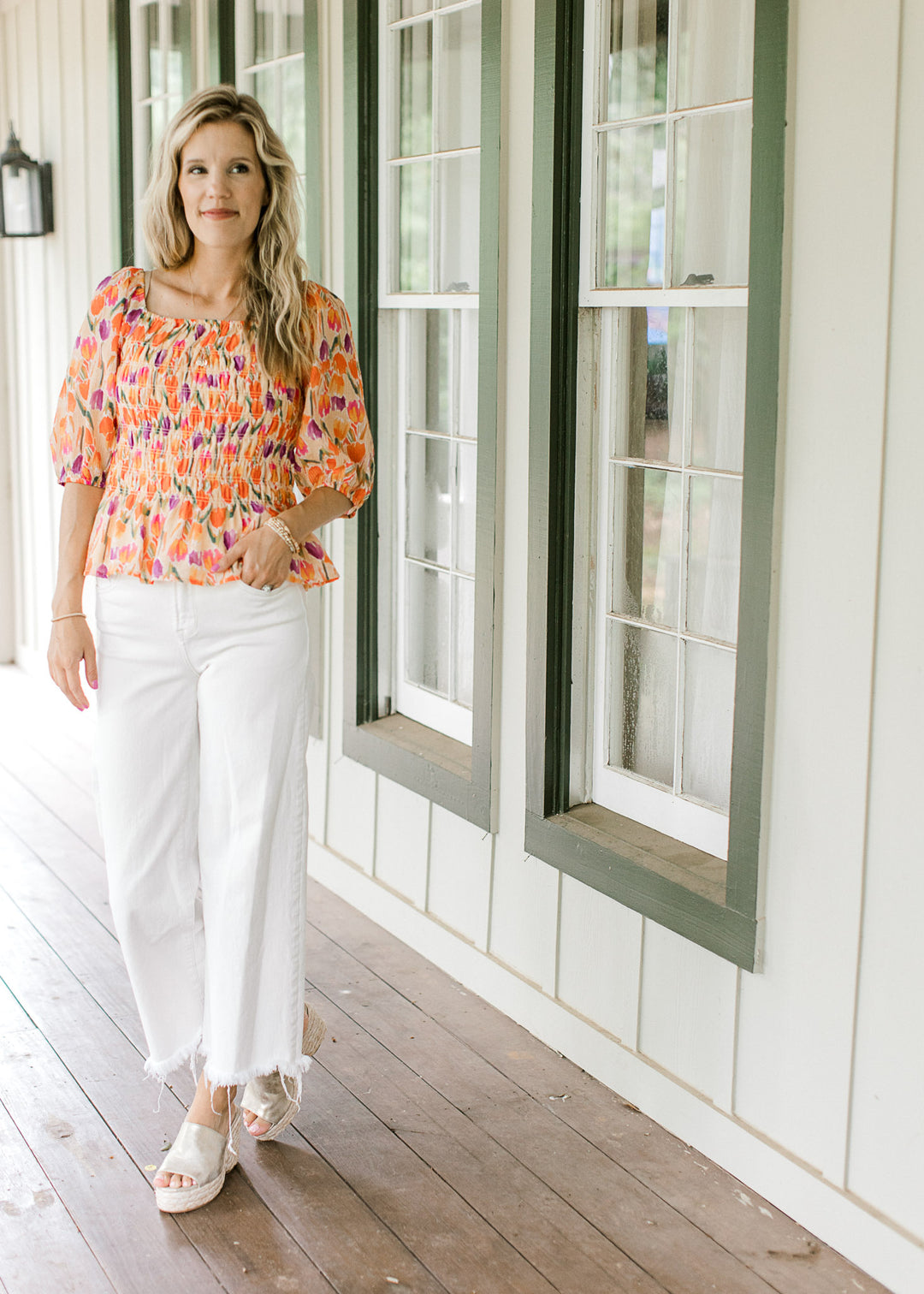 Model wearing white jeans and wedges with a peach top with purple, gold and green tulips. 