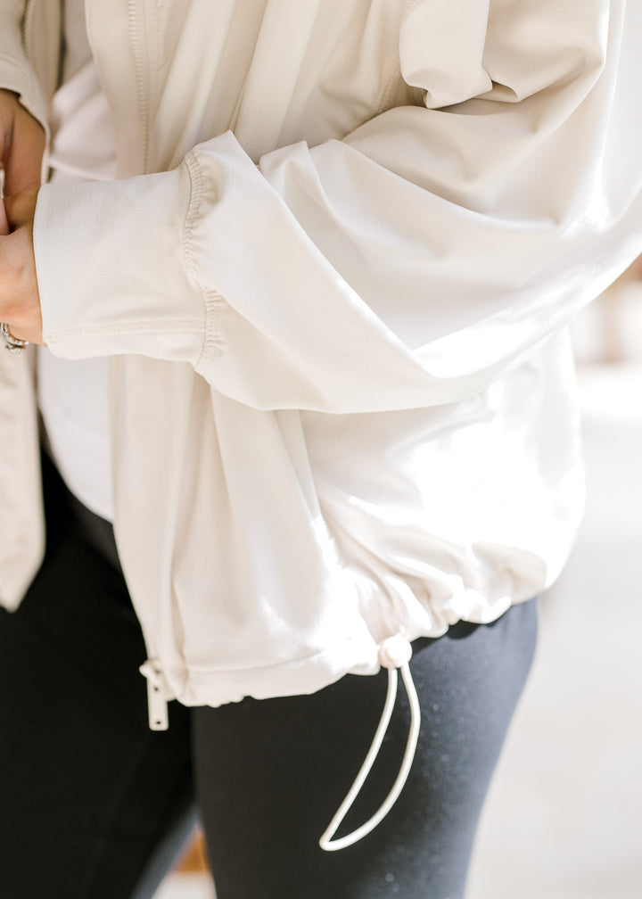 Close up of bungee cord along the bottom of a full zip cream jacket with long sleeves. 