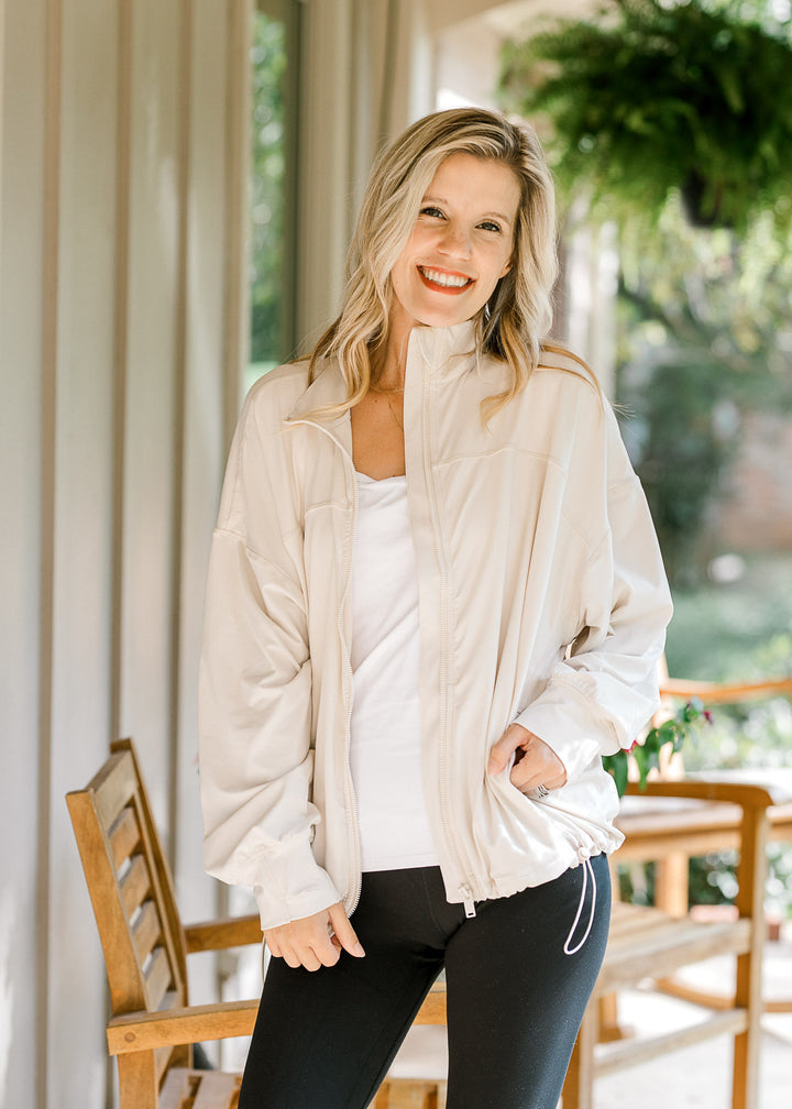 Model wearing a cream jacket with long sleeve with cuff and thumb hole, zipper and bungee bottom.