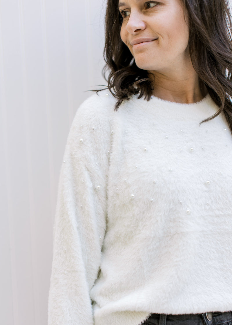 Model wearing an ivory sweater with pearl embellishments, round neck and long sleeves. 