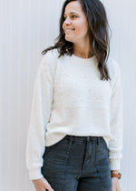 Model wearing jeans with an ivory sweater with pearl embellishments, round neck and long sleeves. 
