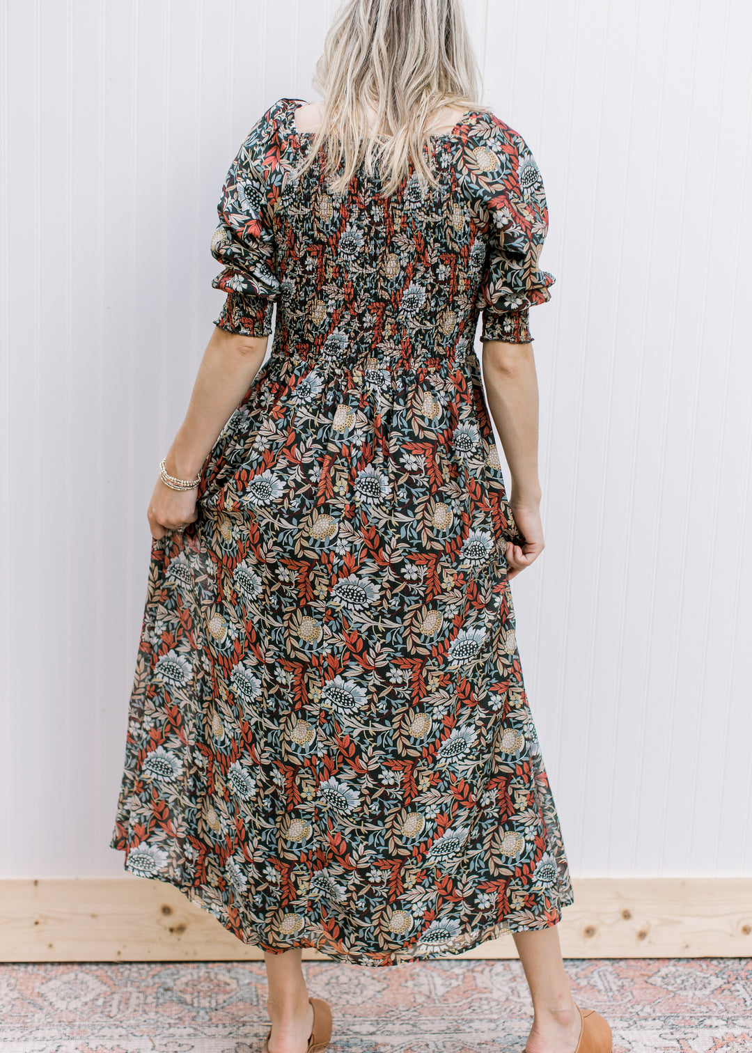 Back view of Model wearing a deep rust short sleeve dress with a floral pattern and smocked bodice.