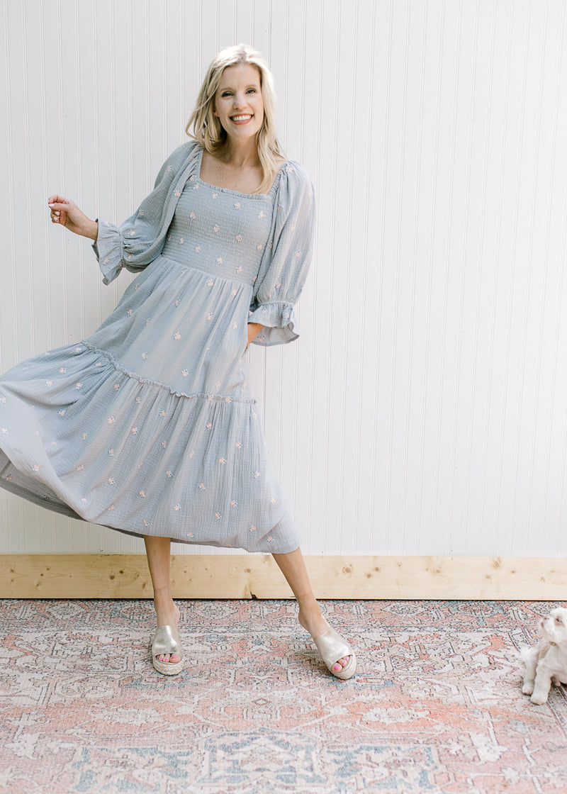 Model wearing a blue dress with pink embroidered flowers, a gauze material and poet 3/4 sleeves.