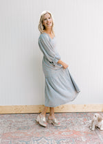 Model wearing a steel blue dress with pink embroidered flowers, smocked bodice and pockets.