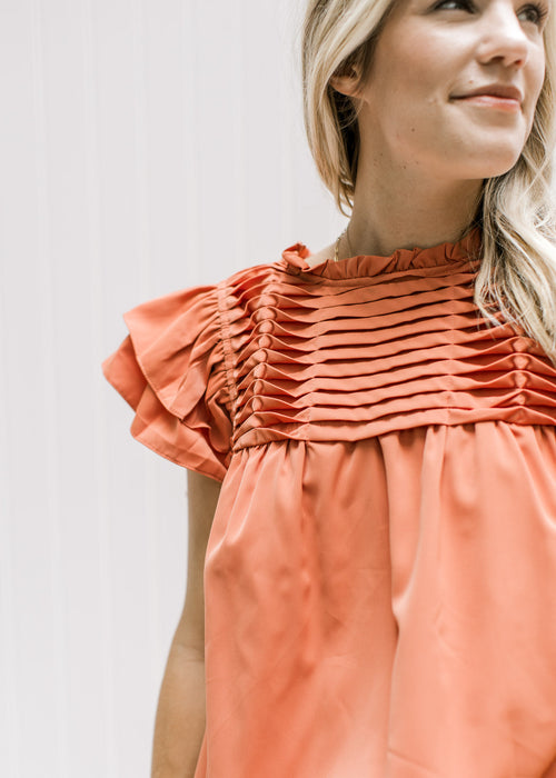 Model wearing a persimmon colored top with a pleated bodice with twisted pleats and short sleeves. 