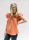 Model wearing a persimmon colored top with a pleated bodice, ruffle neck and ruffle short sleeves. 