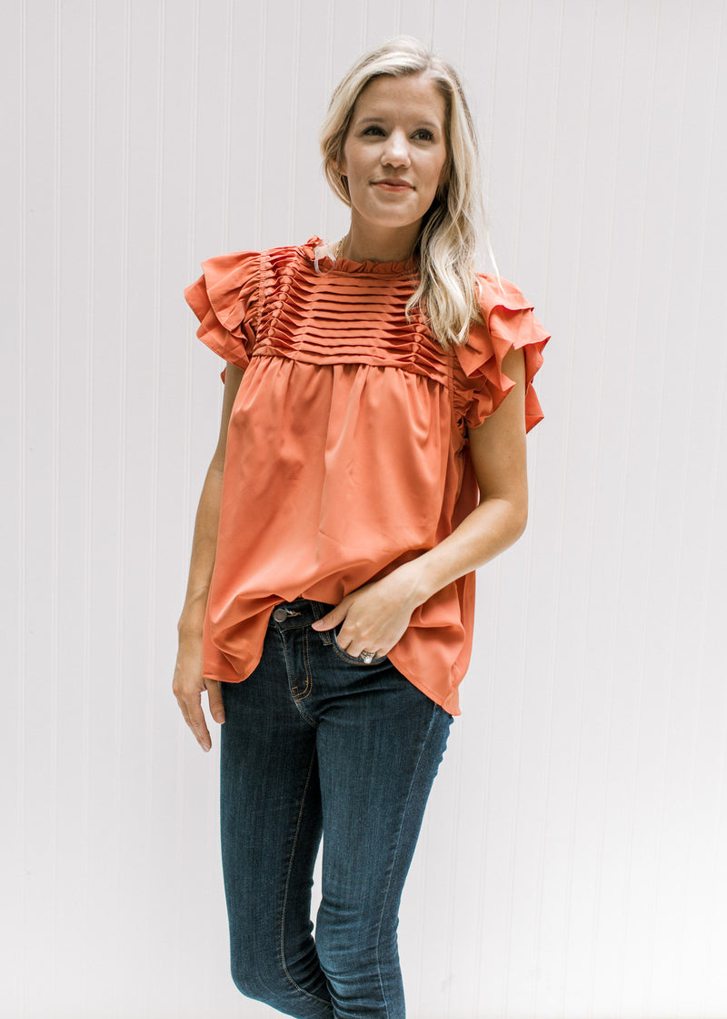 Model wearing jeans with a persimmon colored top with a pleated bodice and ruffle short sleeves. 