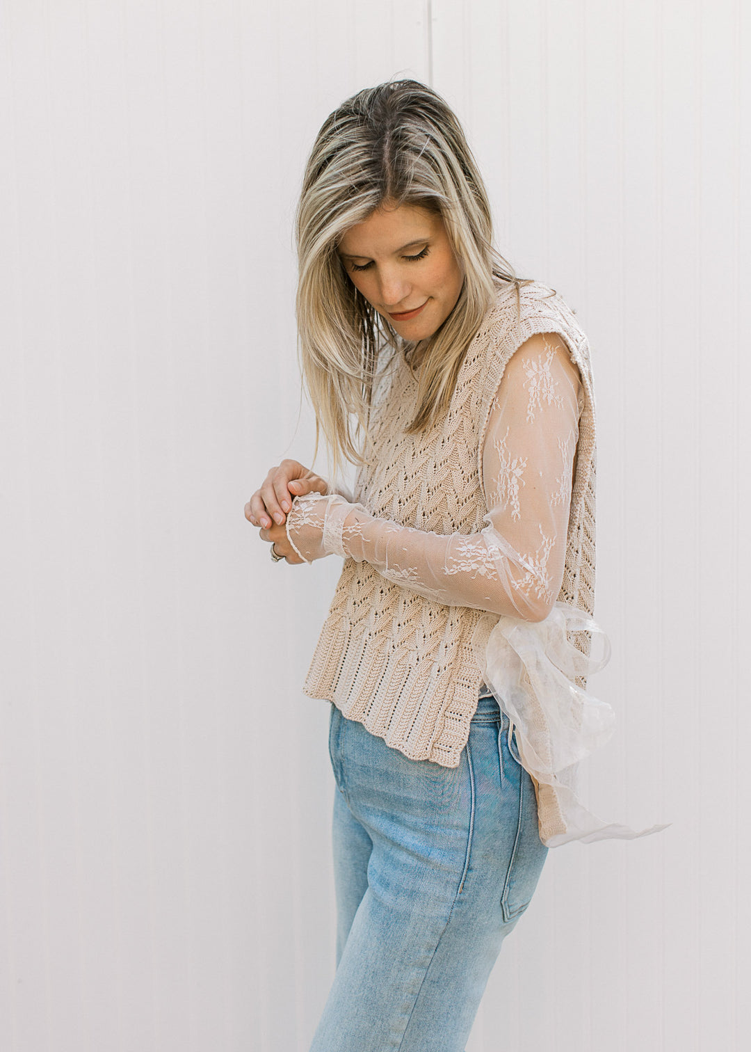 Model wearing sheer floral lace top under an oatmeal sleeveless v-neck with ribbon side tie. 