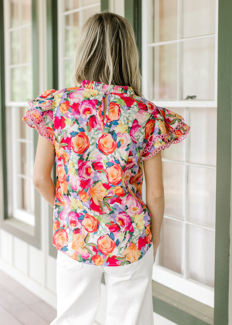 Back view of Model wearing a bright floral top with flutter short sleeves and ruffle neck.