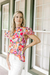 Model wearing white jeans with a bright floral top with flutter short sleeves and ruffle neck.