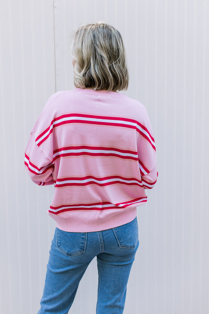 Back view of Model wearing a pink sweater with red and white stripes, round neck and long sleeves.