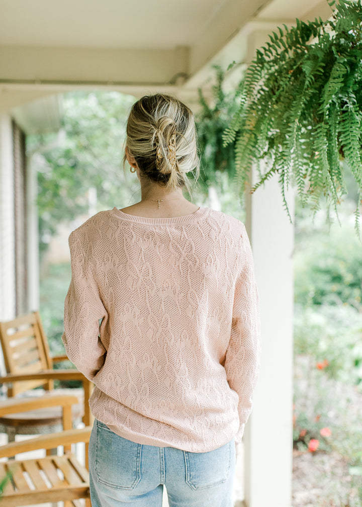 Back view of Model wearing a blush sweater with loose knit, floral design and long sleeves. 