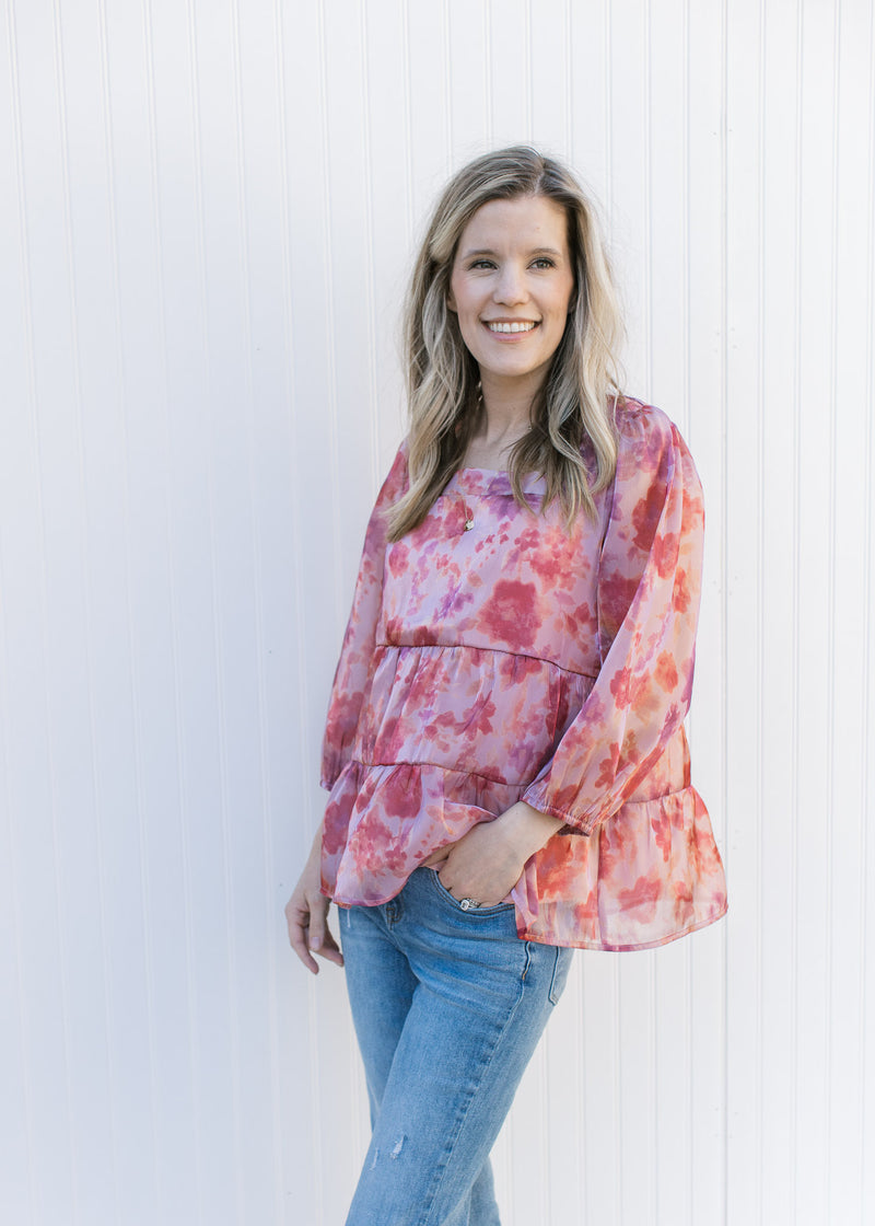 Model wearing jeans with a pink top with a blurred floral pattern, square neck and sheer sleeves.