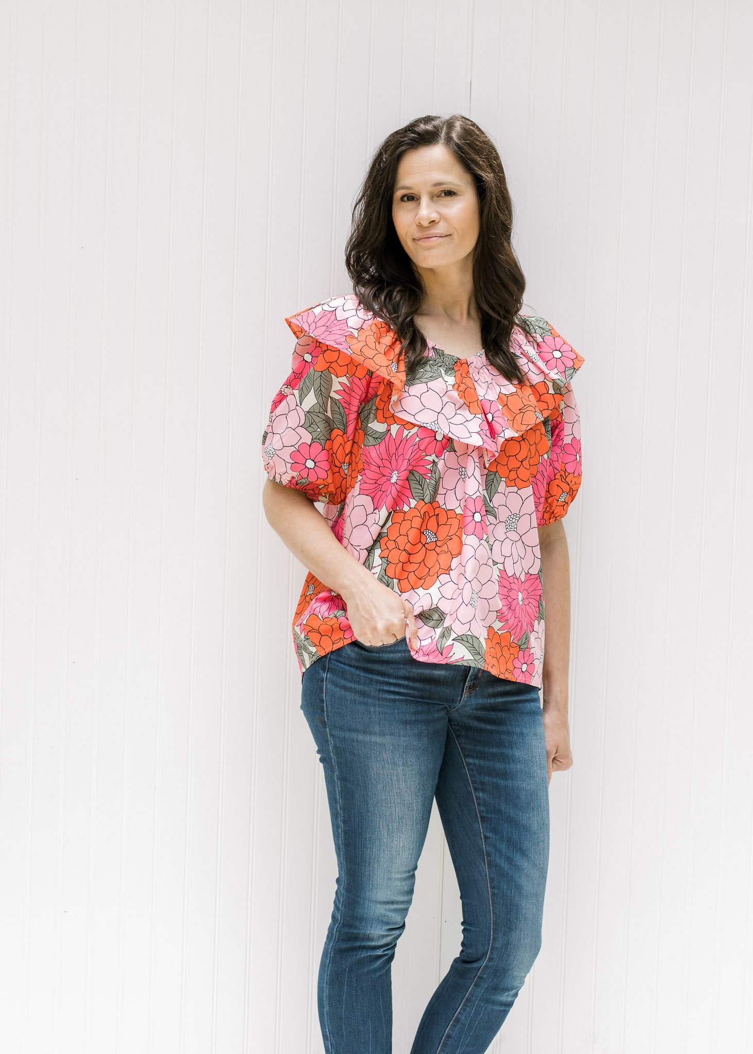 Model wearing jeans with a pink floral top with bubble short sleeves and a boat neck with a ruffle. 