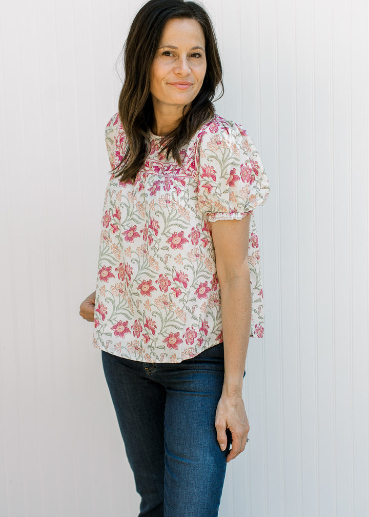 Model wearing a cream top with pink, yellow and green floral a round neck and bubble short sleeves.
