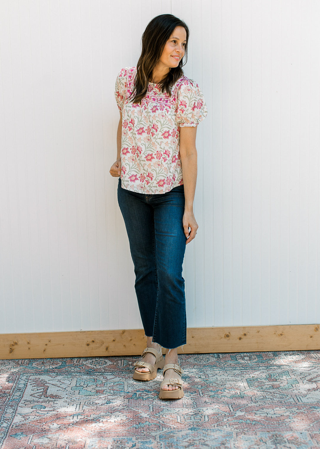 Model wearing jeans, sandals and a cream short sleeve top with pink, yellow and green floral. 