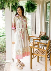 Model wearing sandals and a cream midi with gold and pink print, short sleeves and a tie waist. 