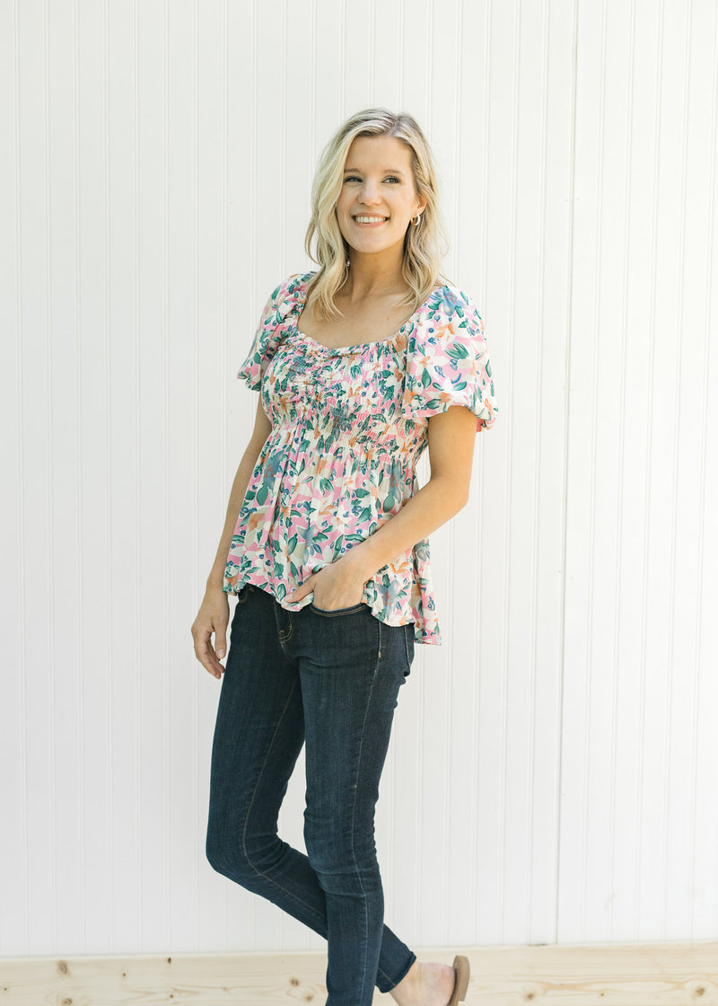 Model wearing jeans and sandals with a pink floral top with short sleeves and a smocked bodice. 
