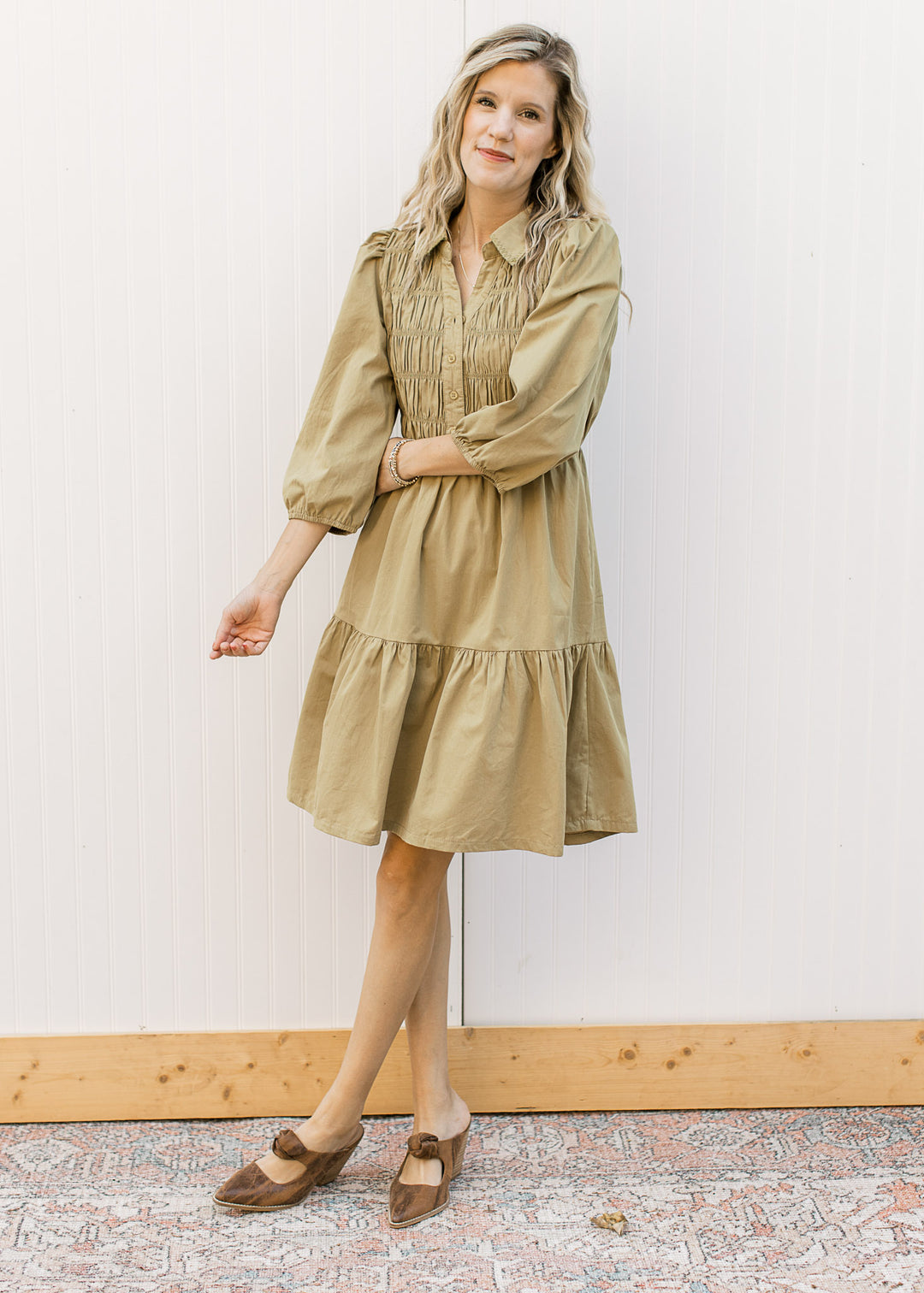 Model wearing heals and a greenish-brown dress with a smocked bodice and 3/4 bubble sleeves.