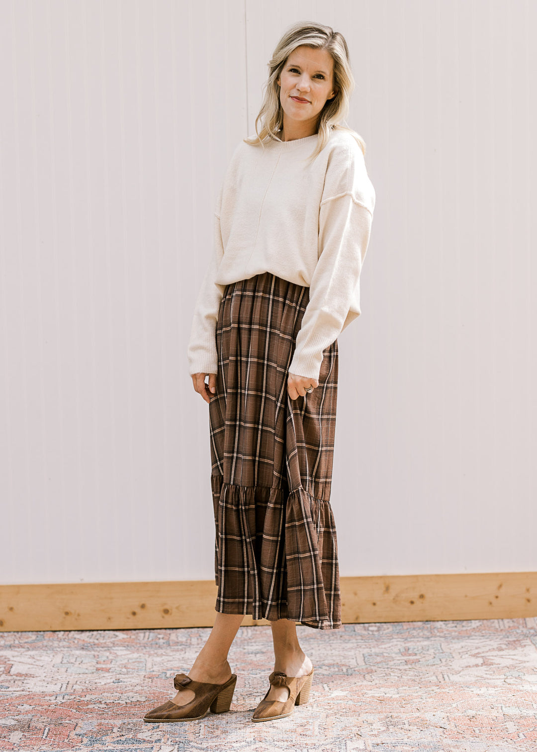 Model wearing a cream long sleeve sweater over a brown and cream plaid midi. 
