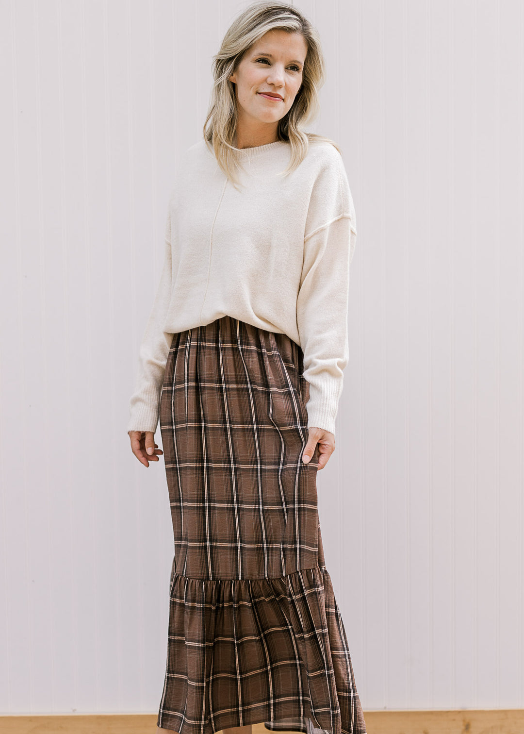 Model wearing a cream long sleeve sweater over a brown, black and cream plaid midi. 