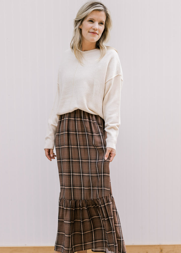 Model wearing a cream long sleeve sweater over a brown, black and cream plaid midi. 