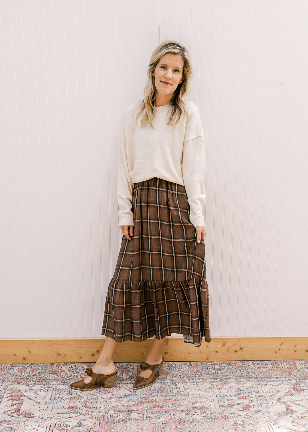 Model wearing a cream long sleeve sweater, heels and a brown, black and cream plaid midi. 