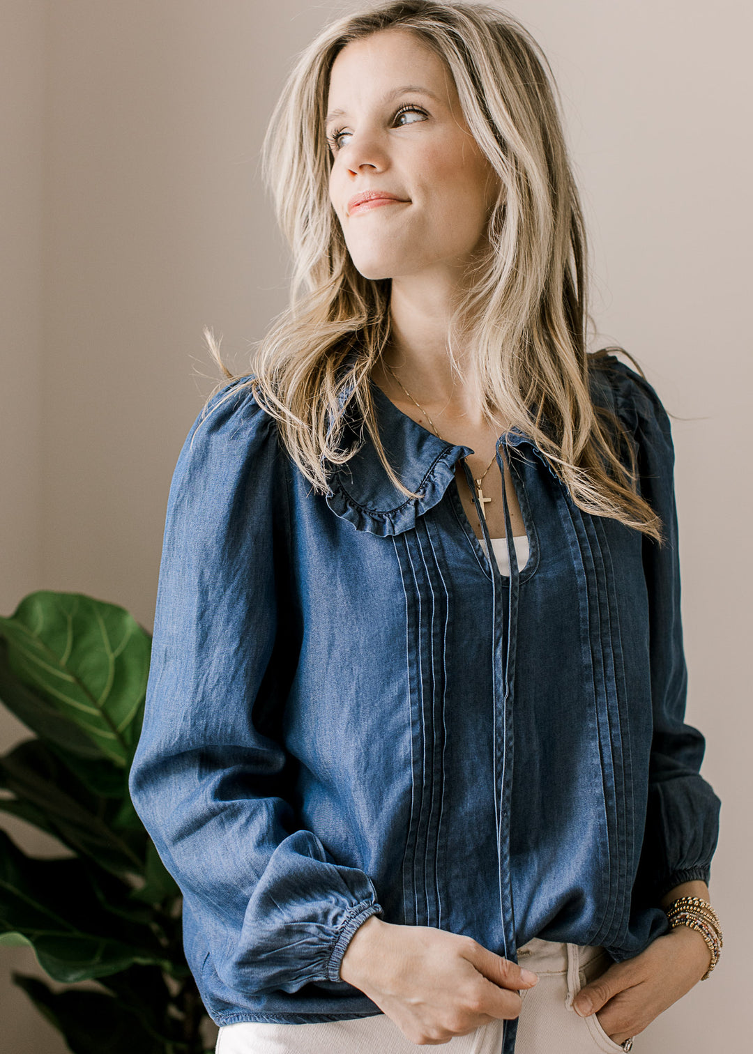 Model wearing a long sleeve blue chambray top with flap collar with ruffle and v-neck with tie. 