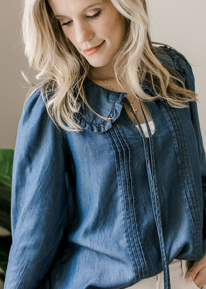Close up of flap collar with a ruffle and tie v-neck on a long sleeve blue top with pleated bodice. 