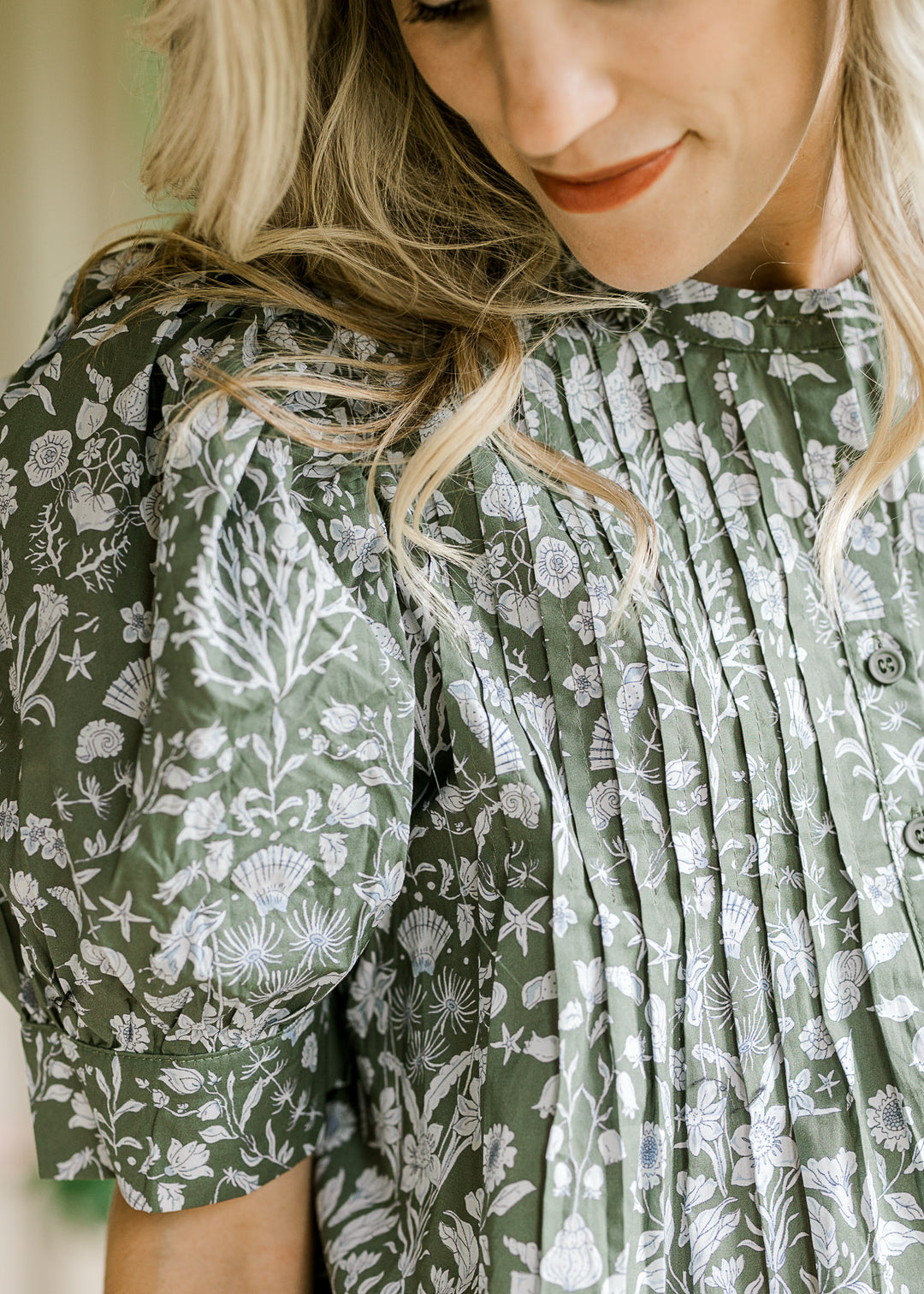 Model wearing an olive top with a blue print, micro pleats on the bodice and short puff sleeves. 