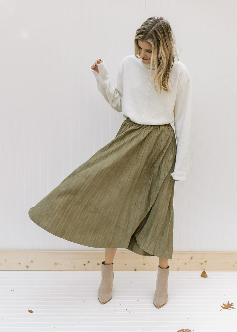 Model wearing a cream top with a  pleated olive skirt with a velvet material and a midi length. 