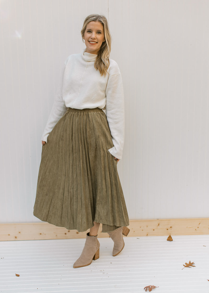 Model wearing a cream top, booties and a pleated olive midi skirt with a velvet material. 
