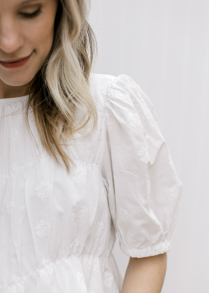 Close up of pleating detail and floral texturing on a white short sleeve top with a round neck. 