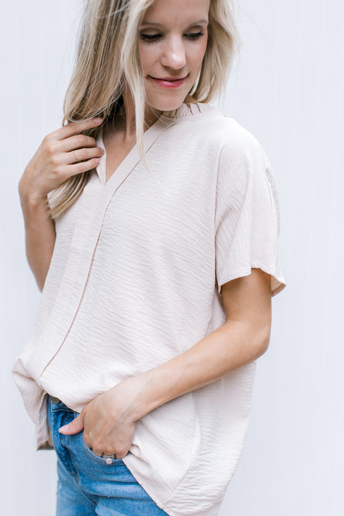 Model wearing an oatmeal colored short sleeve top with a v-neck and a pleat down the center.