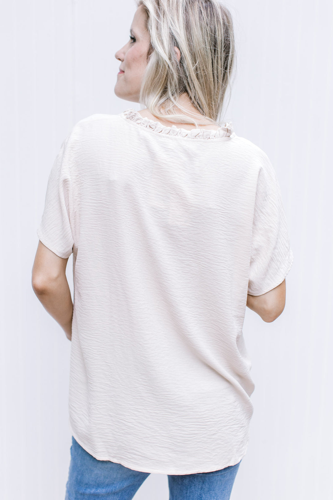 Back view of Model wearing an oatmeal colored short sleeve top with a ruffle at the neck. 